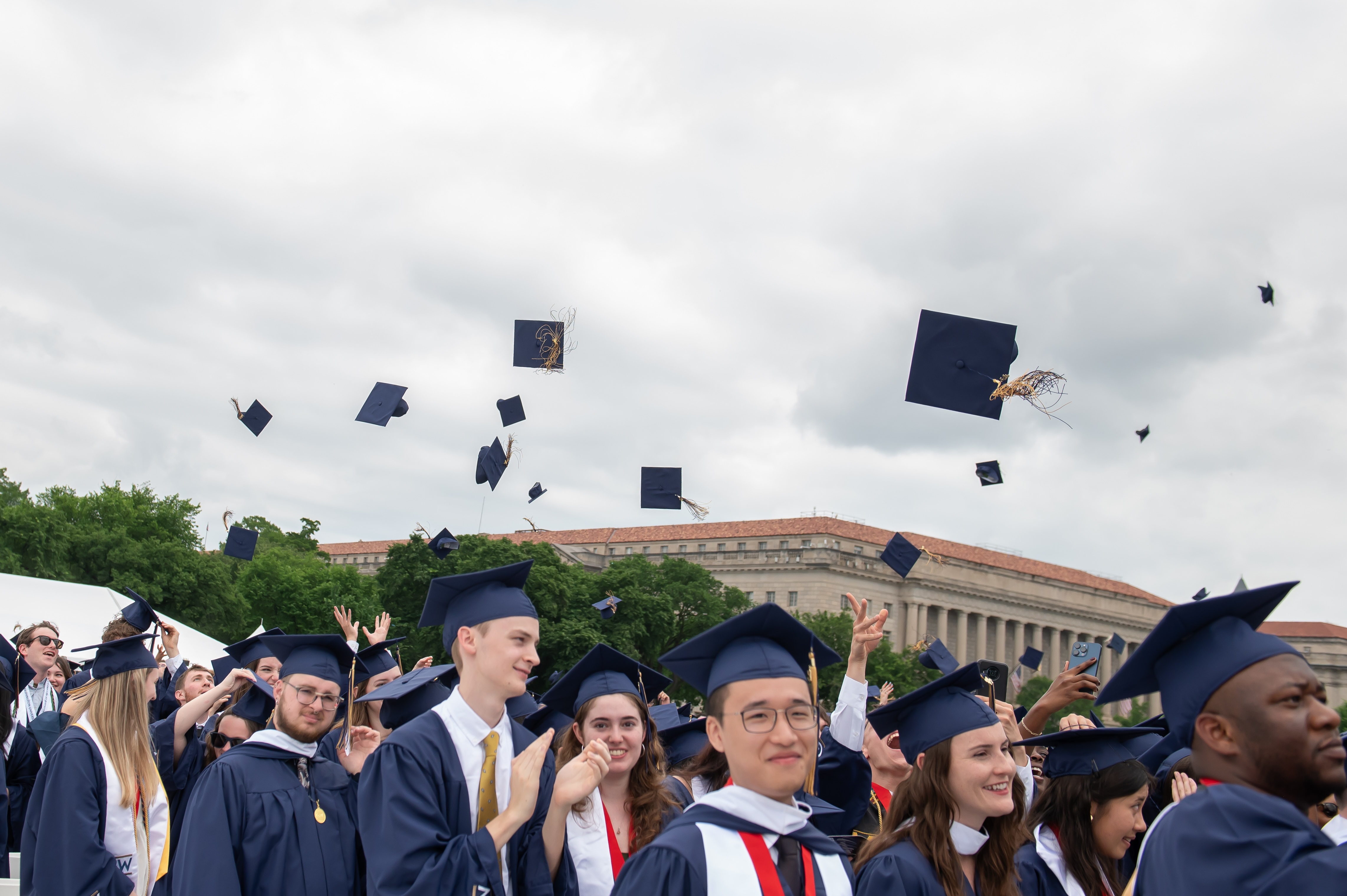 The George Washington University graduates celebrating commencement by throwing their mortar boards in the air.  Building and trees are in the background.