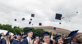 The George Washington University graduates celebrating commencement by throwing their mortar boards in the air.  Building and trees are in the background.